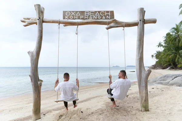 Achterzijde van een homoseksueel koppel schommelend op een schommel in een tropisch strand — Stockfoto