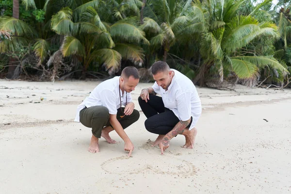 Omosessuale maschio coppia disegno un cuore in il sabbia su un tropicale spiaggia — Foto Stock