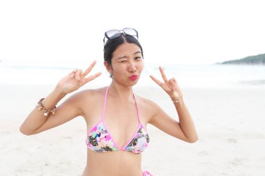 Asyalı kadın kumsalda göz kırparken parmaklarıyla zafer işareti yapıyor.