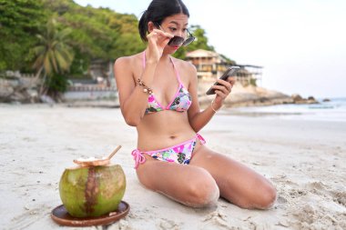 Sahilde cep telefonu ekranını okumak için gözlüğünü çıkarıyor.