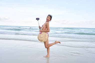 Asyalı flamalı komik poz veriyor ve plajda cep telefonuyla selfie çubuğu tutuyor.
