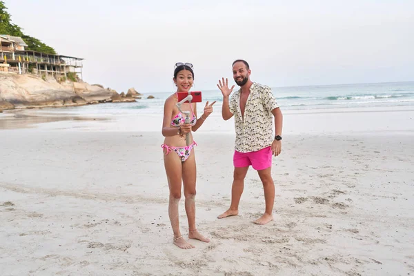 Streamer in costume da bagno registrazione in diretta mentre un uomo saluta la fotocamera sulla spiaggia — Foto Stock
