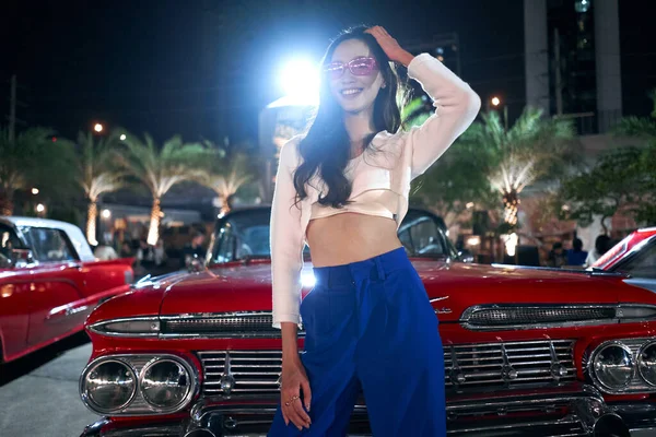 Sensuele aziatische vrouw poseren in de voorkant van een luxe antieke auto in een nacht kermis — Stockfoto