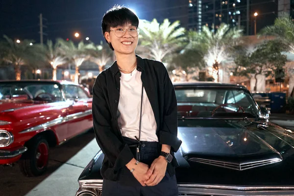 Uśmiechnięta niebinarna osoba opierająca się o luksusowy samochód na nocnych targach — Zdjęcie stockowe