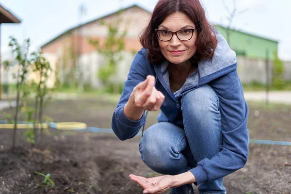 Crouching caucasian woman showing a seed while looking at camera in a vegetable home garden