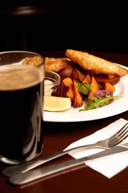 battered fish on a plate with chips on a wooden table in pub and pint of stout. unhealthy food
