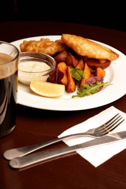 battered fish on a plate with chips on a wooden table in pub and pint of stout. unhealthy food