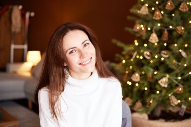 Attractive millennial female sitting in chair in decorated living room. Christmas tree with decoration on background