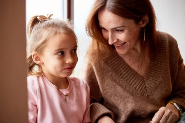 Little caucasian girl with mother unpacking gift boxes at home near window. Happy family christmas concept