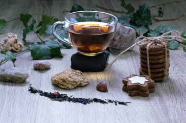 A cup of black tea made from a large leaf stands on the stone. There are biscuits, grains of tea, stones and leafs nearby. Light on the cup