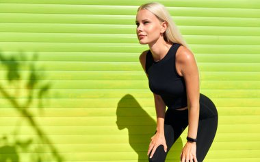 Güneşli bir günde, açık havada idmandan sonra yeşil duvarın yanında dinlenen sarışın bir kadının yatay görüntüsü. Spor salonundaki zayıf kız sabah antrenmandan sonra dinleniyor..