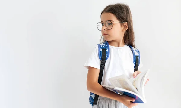 スタジオ水平画像の小さな女の子身に着けています制服 透明メガネは脇に本を読んでいる間に見て あなたのテキストのためのコピースペースと白の背景に対してポーズ — ストック写真
