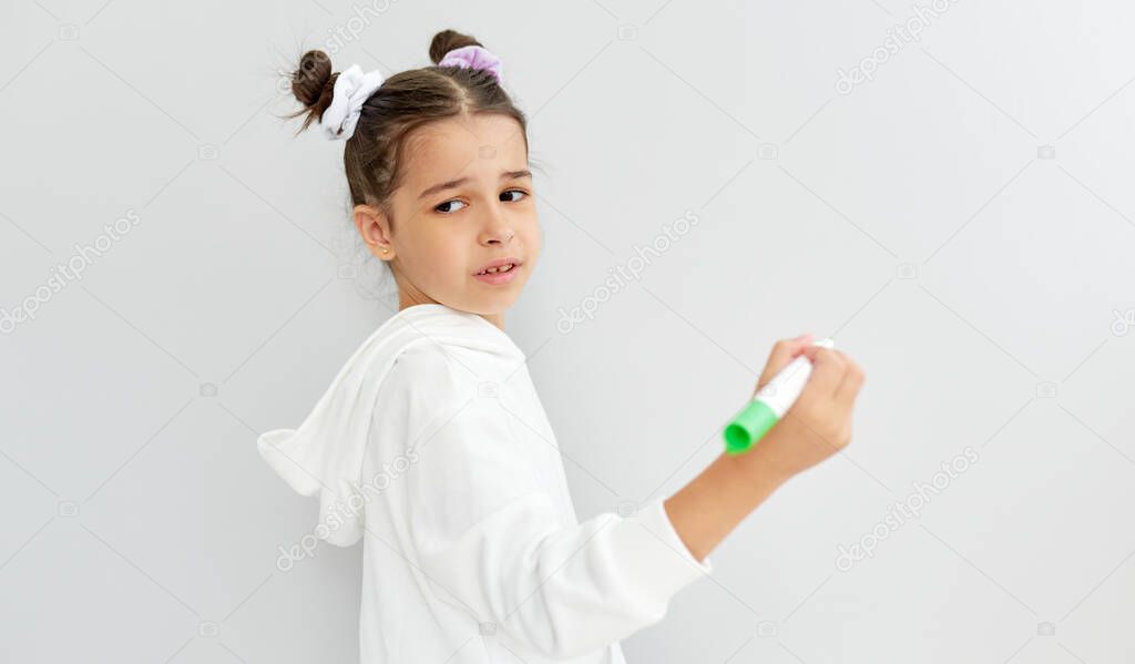 Horizontal image of sad schoolgirl trying to solve something on the whiteboard. An unhappy little girl wearing a white hoodie looking at at the camera during write with green marker on a white board. 