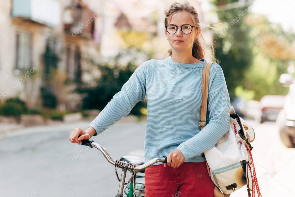 Outdoor image of a student female going to the university with a bike on a sunny day. Pretty woman in casual outfit takes a rest outside after cycling.