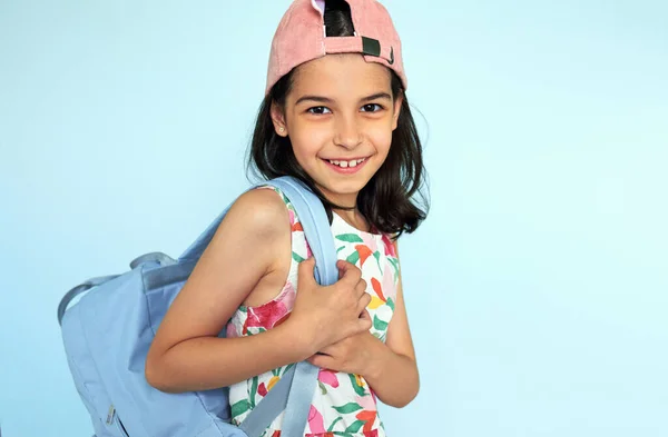 快乐的孩子穿着五颜六色的衣服 戴着粉色的帽子 背着蓝色的背包 看着镜头在蓝色的背景下摆出一副笑脸 带着背包的积极向上的女孩很高兴又回到了夏令营 — 图库照片