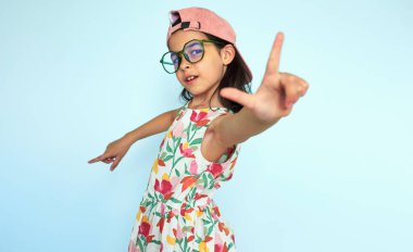 Yazlık elbiseli ve pembe şapkalı çocuk kamerayı parmaklarıyla göstererek eğleniyor. Kameraya parmağını çeviren küçük kız fotoğraf çekiyormuş gibi yapıyor..