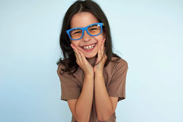 茶色のTシャツを着た陽気な女の子のスタジオの肖像画 そして青い眼鏡 笑顔と楽しい気分は青い背景に隔離された彼女の顔に手を保持しています — ストック写真