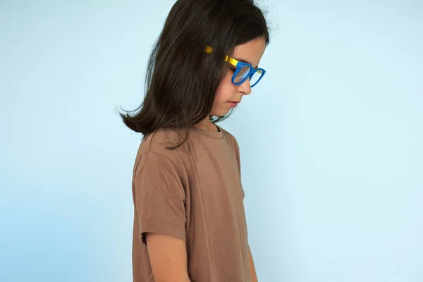 Side view portrait of a cute little girl in brown t shirt and blue and yellow eyeglasses looking at one side posing over blue studio background.