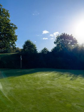 Mavi gökyüzü ve bayrakla yeşil golf sahası, çiğ taneli sonbahar günü