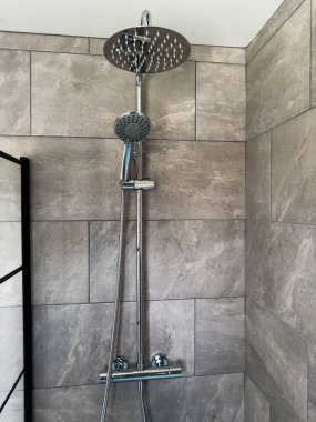 Karanlık lavabosu ve aynası olan küçük modern bir banyo. Krom duşlu ve musluklu gri duvar fayansları. Duş perdesinde siyah ayrıntı