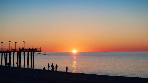 Barevný přímořský plášť, západ slunce a červená obloha. Pláž a molo s lidmi silueta v podsvícení, pobřežní slunce na vodě — Stock fotografie