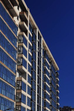 Bir apartmanın ön cephesinin mimari parçası, tuğla duvarlar ve mavi gökyüzüne karşı camdan balkonlar..