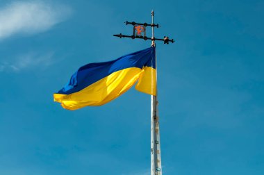 Mavi gökyüzünde Ukrayna bayrağı olan metal kule ve Lviv arması olan rüzgar gülü. Sarı ve mavi bayrak rüzgarda.