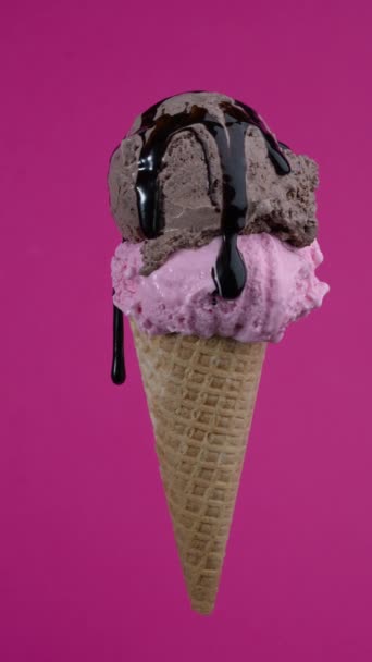 Tasty ice cream cone with two chocolate and strawberry flavors scoops. Pouring syrup topping in waffle cone on pink background. Soft cream, gelato icecream scoop in waffle cone over colorful wallpaper — Stockvideo