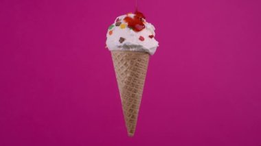 Waffle külahında vanilyalı dondurma ve çilekli sos. Renkli süslemelerle süslenmiş tatlı, dönen yumuşak krema, pembe arka planda dondurma kepçesi