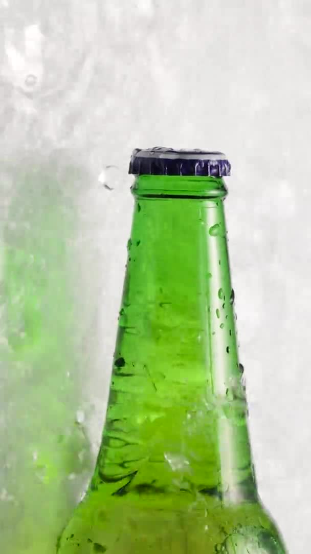 Groene glazen fles koud bier koel op ijsblokjes, lage hoek, zijaanzicht, waterdruppels op glas, verfrissende bevroren alcoholische drank met spetters over ijsachtergrond close-up in slow motion — Stockvideo