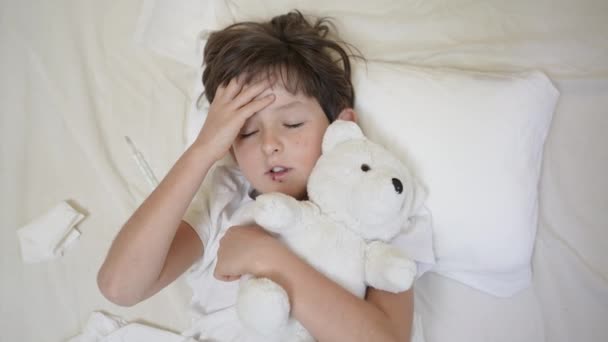 熱のある病気の子供の男の子はテディベアとベッドの枕の上に横たわっています。エネルギー就学前の子供の疲れ不足、温度、発熱インフルエンザの症状と治療、子供医療、コロナウイルスの症状 — ストック動画