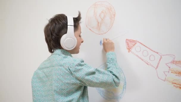 Nette lustige Schulkind Junge Künstler in Kopfhörer Zeichnung Malerei Bild mit Kreide, Wachsmalstift auf weißer Wand, fokussierte intelligente Kind genießen kreative Kunst Hobby-Aktivität zu Hause, Kinder Entwicklungskonzept — Stockvideo
