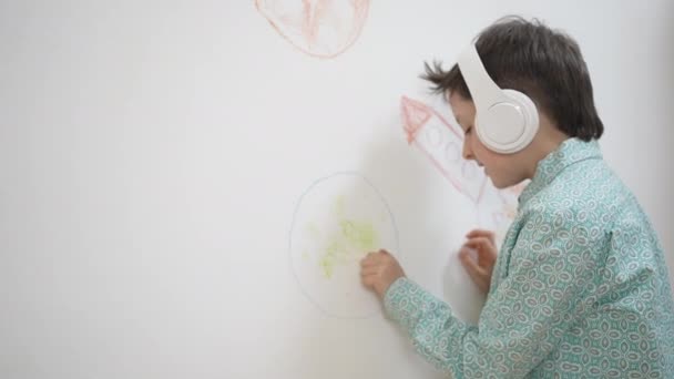 Nette lustige Schulkind Junge Künstler in Kopfhörer Zeichnung Malerei Bild mit Kreide, Wachsmalstift auf weißer Wand, fokussierte intelligente Kind genießen kreative Kunst Hobby-Aktivität zu Hause, Kinder Entwicklungskonzept — Stockvideo