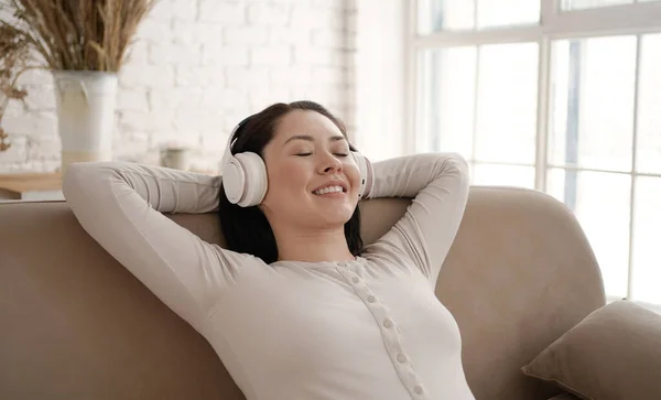 Junge Asiatin entspannt auf bequemem Sofa mit geschlossenen Augen und trägt Kopfhörer. Hübsche Mischlingshündin genießt das Hören von Chill-Musik Audio-Sound Meditation Gefühl kein Stress zu Hause. — Stockfoto