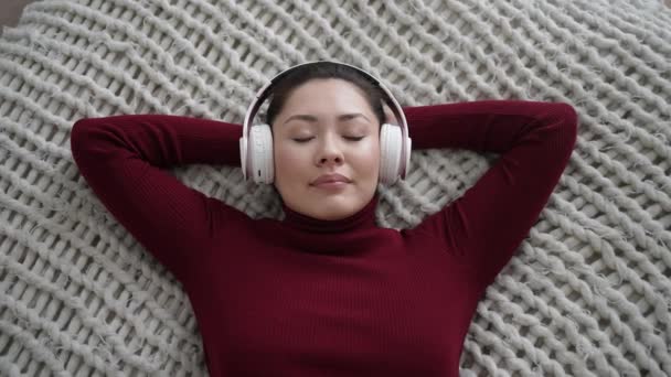 Молода багатонаціональна азіатська жінка розслабляється на зручному дивані з закритими очима в навушниках. Досить змішана раса леді насолоджується прослуховуванням холодної музики звуку, медитуючи, не відчуваючи стресу вдома . — стокове відео