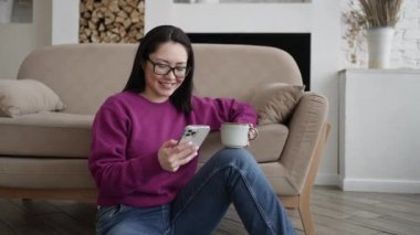 Genç Asyalı kadın sosyal medyada akıllı telefon sörfü yapıyor, haberleri kontrol ediyor, mobil oyunlar oynuyor ya da koltukta oturmuş mesaj yazıyor. Karışık ırk hanımı evinde cep telefonu teknolojisiyle vakit geçiriyor..