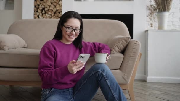 Jovem asiática usando telefone inteligente surfar redes sociais, verificar notícias, jogar jogos móveis ou mensagens de texto sentado no sofá. Misturado corrida senhora passar tempo em casa com a tecnologia gadget celular. — Vídeo de Stock