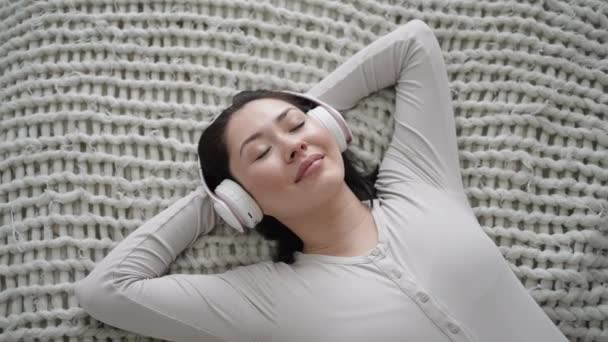 Junge multiethnische Frau chillt auf bequemem Sofa mit geschlossenen Augen mit drahtlosen Kopfhörern, asiatische Mädchen genießen Chill-Musik und Audio-Sound entspannt zu Hause hören. Draufsicht in Zeitlupe — Stockvideo