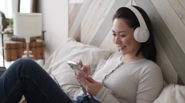 Genç Asyalı kadın sosyal medyada akıllı telefon sörfü yapıyor, haberleri kontrol ediyor, mobil oyunlar oynuyor ya da koltukta oturmuş mesaj yazıyor. Karışık ırk hanımı evinde cep telefonu teknolojisiyle vakit geçiriyor..