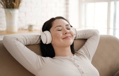 Genç Asyalı kadın rahat bir kanepede, kulaklık takıp gözleri kapalı dinleniyor. Oldukça karışık ırk hanımı, rahat müzik dinlemeyi sever. Sesli meditasyon yapar. Evde stres hissetmez..