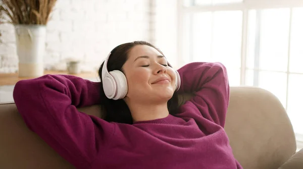 Genç Asyalı kadın rahat bir kanepede, kulaklık takıp gözleri kapalı dinleniyor. Oldukça karışık ırk hanımı, rahat müzik dinlemeyi sever. Sesli meditasyon yapar. Evde stres hissetmez..