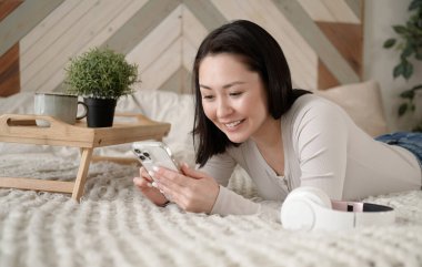 Genç Asyalı kadın sosyal medyada akıllı telefon sörfü yapıyor, haberleri kontrol ediyor, mobil oyunlar oynuyor ya da koltukta oturmuş mesaj yazıyor. Karışık ırk hanımı evinde cep telefonu teknolojisiyle vakit geçiriyor..
