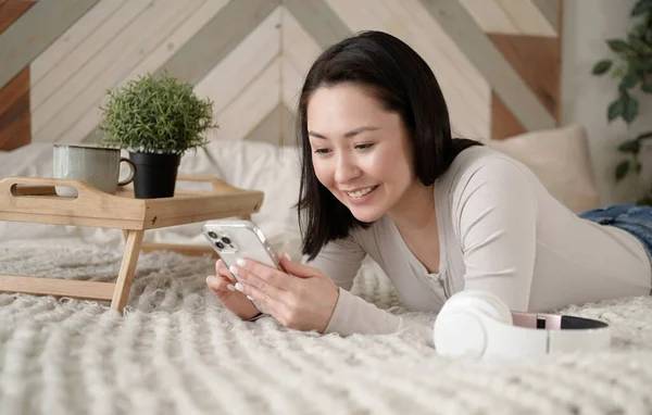 Wanita asia muda menggunakan telepon pintar berselancar di media sosial, memeriksa berita, bermain game mobile atau mengirim pesan duduk di sofa. Wanita ras campuran menghabiskan waktu di rumah dengan teknologi gadget sel. Stok Foto Bebas Royalti