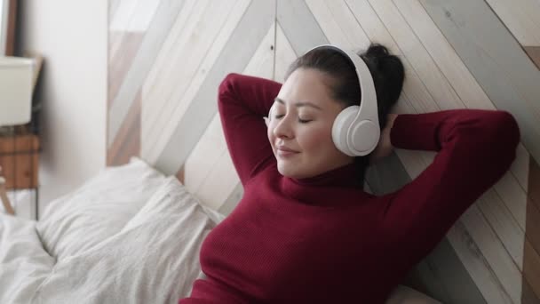年轻迷人的亚洲女人头戴耳机，紧闭双眼，在舒适的沙发上放松。非常健康镇定的女孩喜欢听冷冰冰的音乐，听音频，冥想，在家里没有压力. — 图库视频影像
