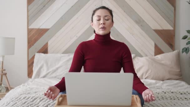 Χαλαρός μικτή φυλή νεαρή ασιατική γυναίκα κάθεται στο κρεβάτι της με διπλωμένα σε χέρια χειρονομία mudra, μειώνοντας το άγχος κατά τη διάρκεια της εργάσιμης ημέρας, ψύξη ή να κάνει ασκήσεις αναπνοής γιόγκα, εργασίας από το σπίτι με φορητό υπολογιστή — Αρχείο Βίντεο