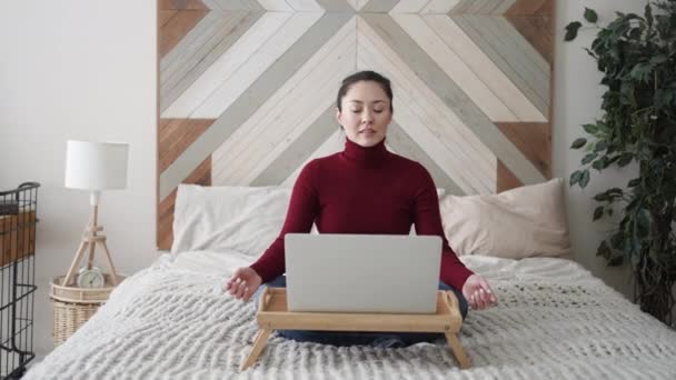 Χαλαρός μικτή φυλή νεαρή ασιατική γυναίκα κάθεται στο κρεβάτι της με διπλωμένα σε χέρια χειρονομία mudra, μειώνοντας το άγχος κατά τη διάρκεια της εργάσιμης ημέρας, ψύξη ή να κάνει ασκήσεις αναπνοής γιόγκα, εργασίας από το σπίτι με φορητό υπολογιστή — Αρχείο Βίντεο