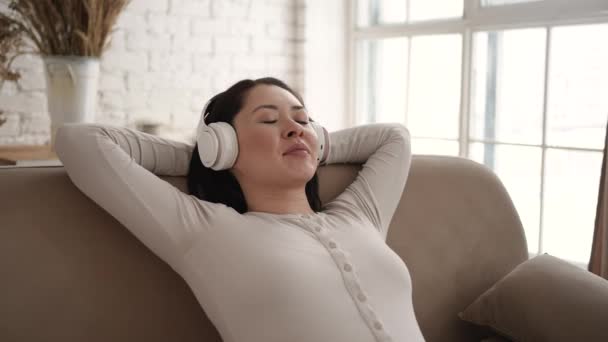 Молода азіатська жінка охолоджує на зручному дивані з закритими очима в бездротових навушниках, змішана расова дівчина насолоджується прослуховуванням холодної музики та звуку, розслаблюючим та медитуючим вдома . — стокове відео