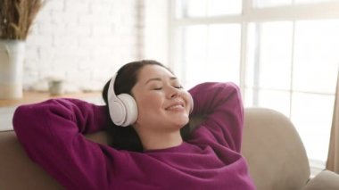 Genç Asyalı etnik kadın rahat bir kanepede, kulaklık takıp gözleri kapalı dinleniyor. Güzel melez ırk kızı, rahat müzik dinlemeyi sever. Sesli meditasyon, evde stres yok..