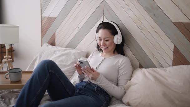 Ung asiatisk etnisk kvinna med hjälp av smarttelefon surfa sociala medier, kontrollera nyheter, spela mobilspel eller sms: a sitter på soffan. Multi-race flicka tillbringa tid hemma med gadget teknik. — Stockvideo