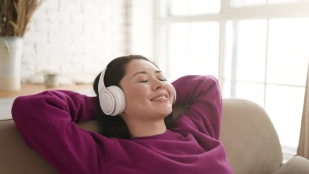 年轻的亚裔妇女戴着耳机，紧闭双眼，坐在舒适的沙发上休息。漂亮的混血儿喜欢听清冷的音乐，听音频，冥想，在家里没有压力. — 图库视频影像